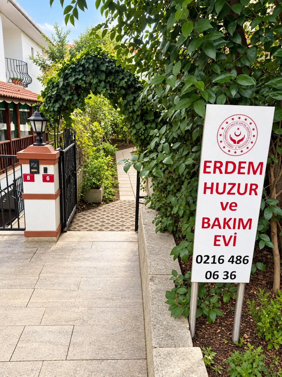 Bahçe girişindeki Erdem Huzur ve Bakım Evi tabelası, telefon numarası ve sarmaşıklı kemer