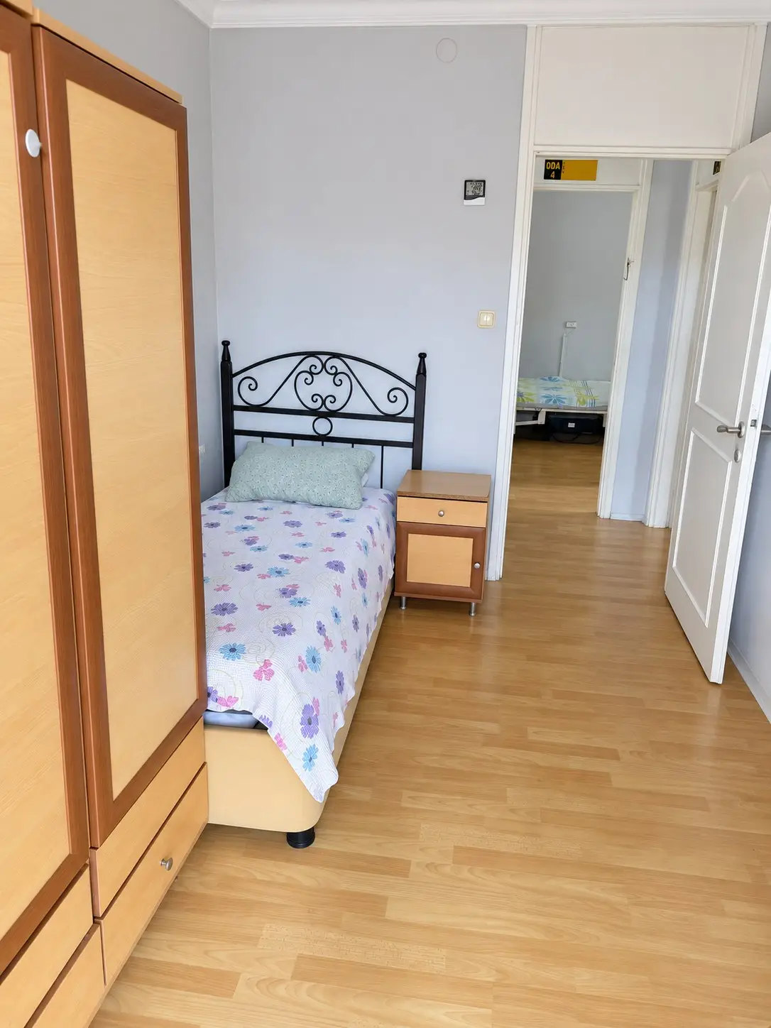 Gardırobun yanından görünen, ferforje başlıklı tek yatak ve komodinli aydınlık oda